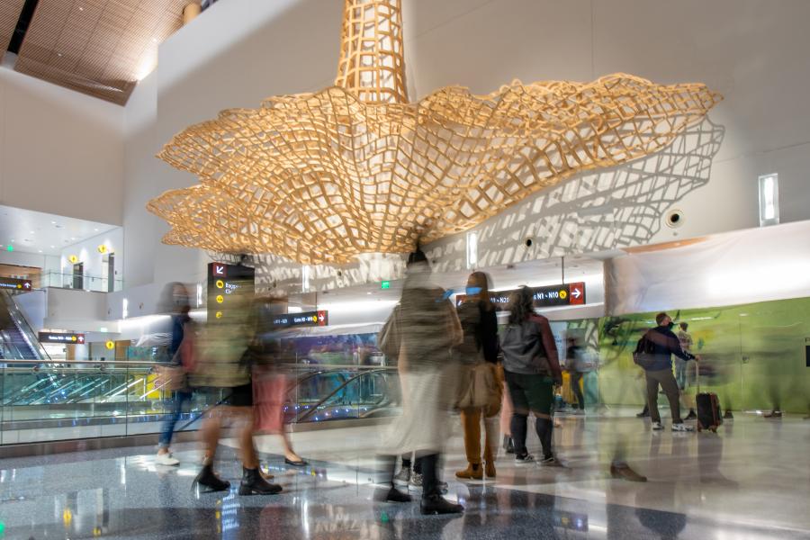 People move through an airport concourse with a large hanging lattice sculpture overhead.