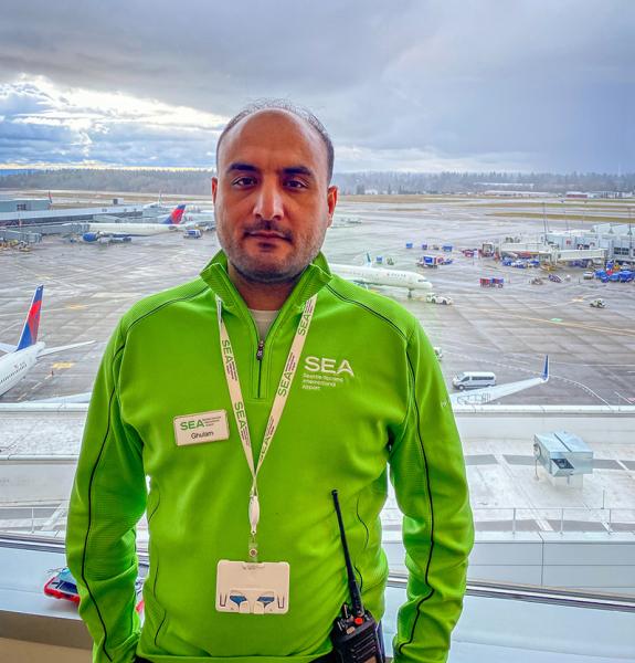 Ghulam Mohmand in a green SEA Airport uniform standing indoors with a view of airplanes and the runway behind them.