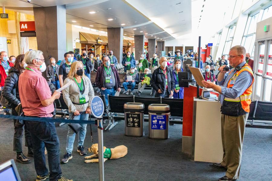 Group of people standing in an airport gate area while listening to an airport staff member who holds a microphone and a clipboard. Most people wear face masks and green lanyards. Rows of seats, signage, and trash and recycling bins are visible, along with a service animal lying on the floor near the front. Large windows line the right side of the space, and gate signs including ‘A4’ are visible in the background.