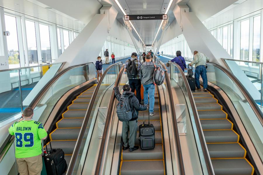 Passengers move through a bright airport corridor using three parallel escalators, with many travelers carrying luggage. Large floor‑to‑ceiling windows line both sides of the space, and overhead signs point toward the train to the airport’s central terminal. The walkway features modern structural supports and clean, open design.