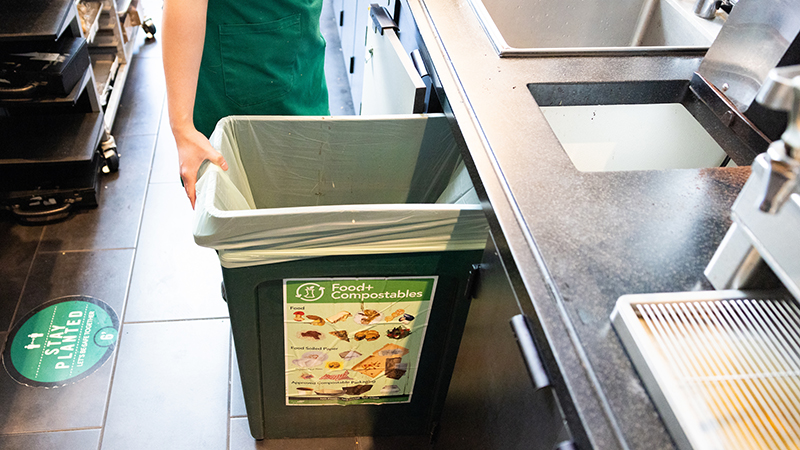 Person holding a food‑scrap compost bin lined with a bag in a commercial kitchen area