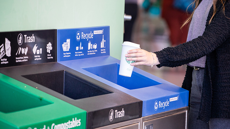 Person placing an empty cup into a recycling bin next to trash and compost bins.