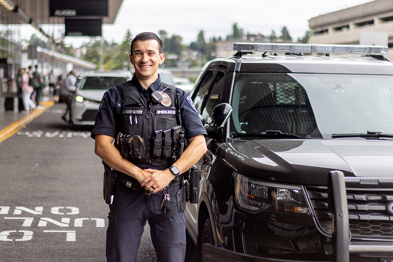A uniformed law enforcement officer stands beside a parked patrol vehicle outside a busy transportation terminal, with people and cars visible in the background.