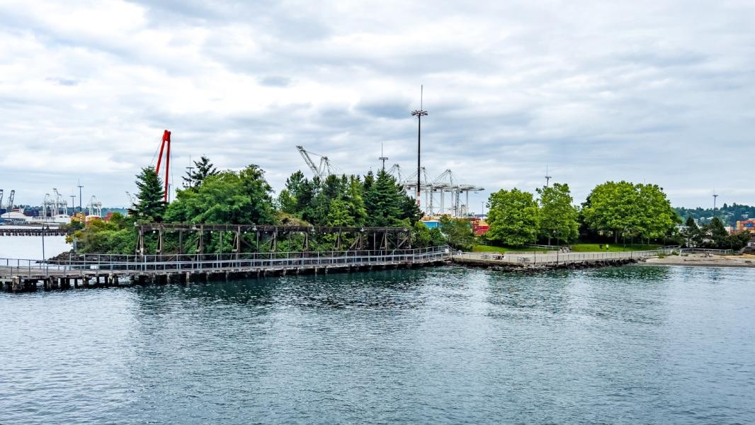 Waterfront shoreline with trees, a wooden walkway, and a small park area, with large industrial cranes and port facilities visible in the background under a cloudy sky.