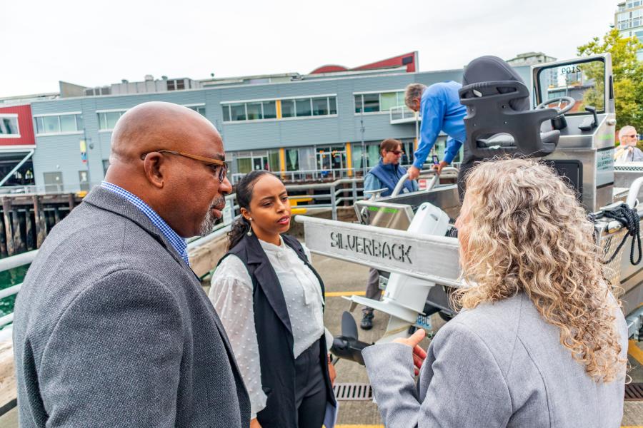 Delmas speaking with two people on a  waterfront dock talking near a small boat, with a building and other people visible in the background.