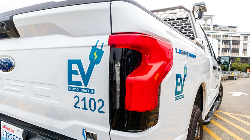 Rear side of a white pickup truck displaying blue ‘EV Port of Seattle’ graphics and the number ‘2102,’ with the truck parked on a paved area near multi‑story buildings.