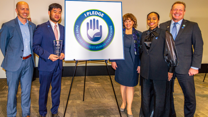 Commissioner Mohamed stands by a sign reading 'I pledge - Port allies against human trafficking'.
