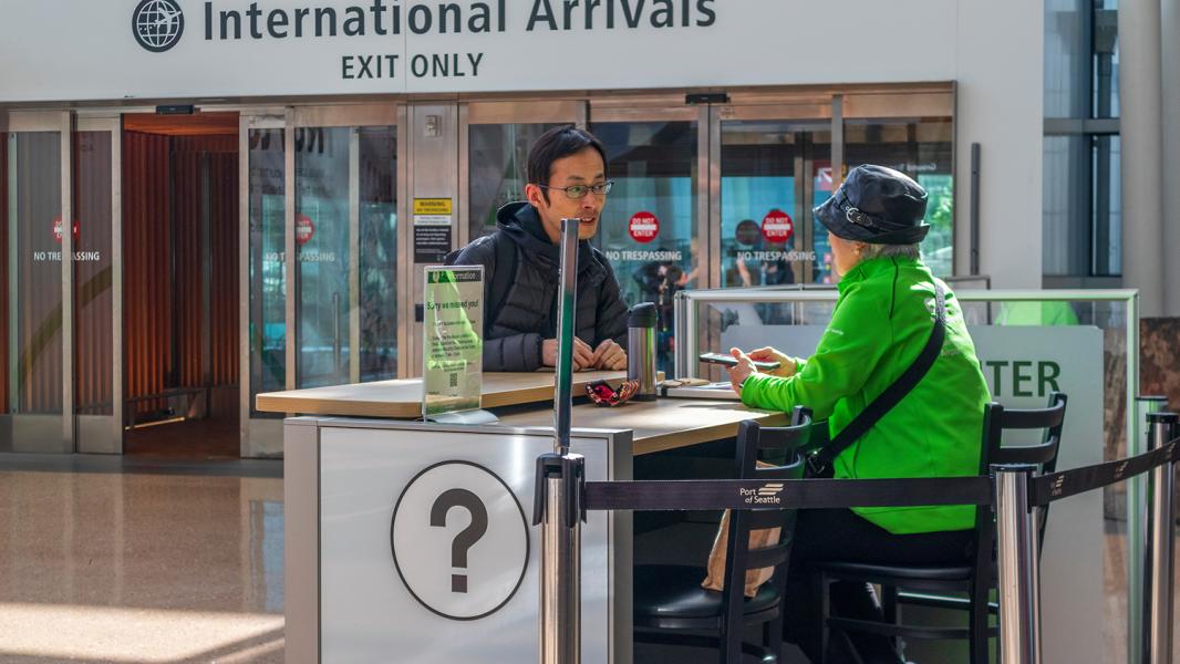 Airport volunteer seated at an information desk talks with a traveler near the International Arrivals exit.