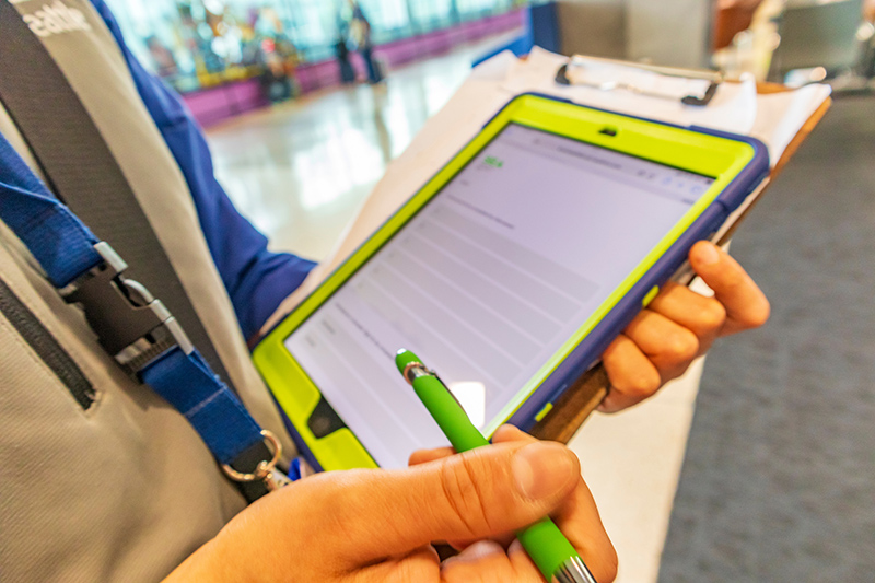 A person holds a tablet on a clipboard and uses a stylus to fill out a form in an indoor public space.