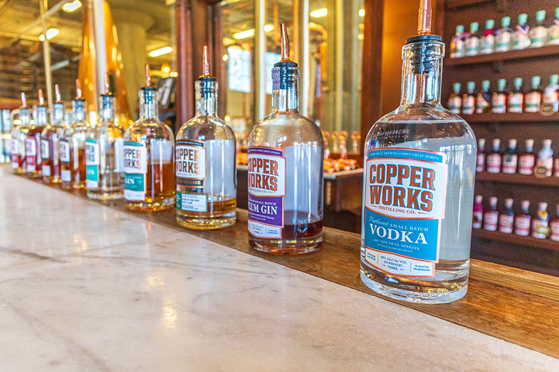 Row of Copperworks distillery bottles on a bar counter with copper stills and shelves in the background.