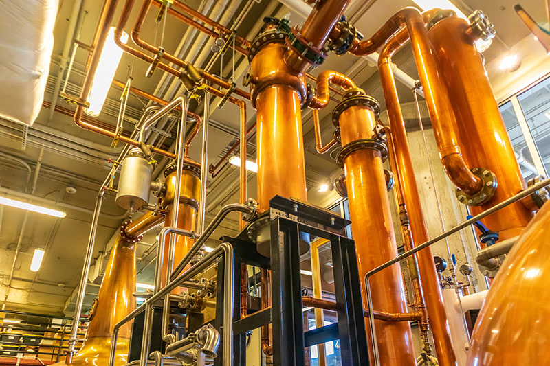 Tall copper distillation columns and pipes inside an industrial distillery.