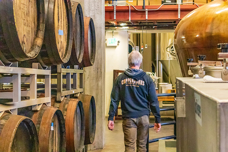 Person walking through a distillery workspace with stacked wooden barrels on one side and large metal equipment on the other.