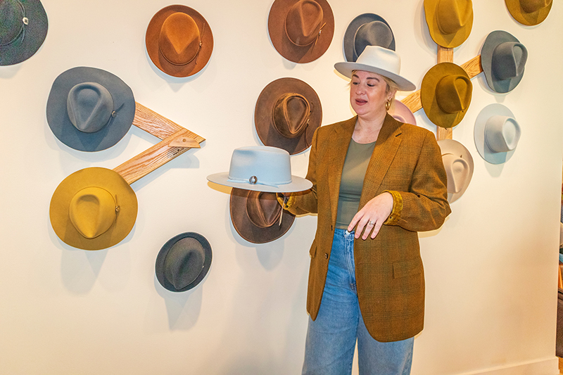 Jamie Slye stands in front of a wall display of hats and holds a light-colored hat in one hand inside a hat shop.