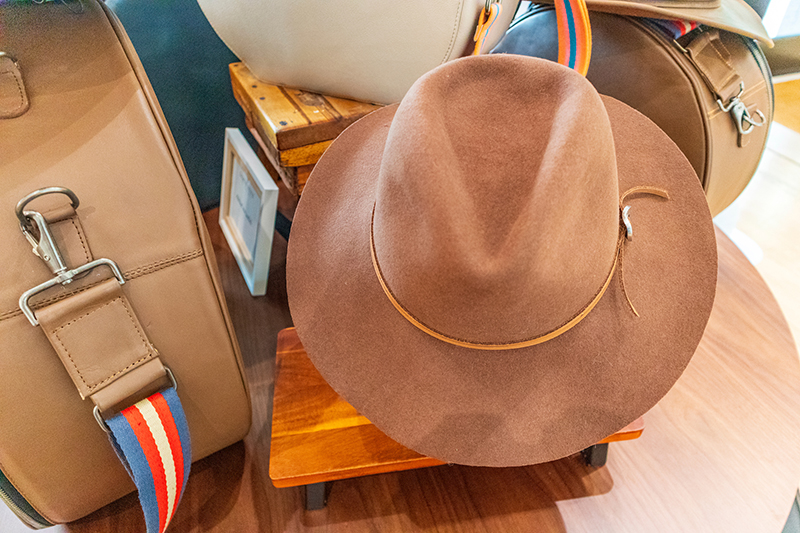 A brown felt hat rests on a small wooden stand among tan bags with striped straps displayed on a table.