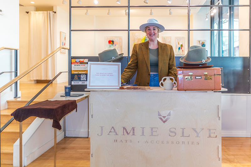 Jamie Slye stands behind a hat shop counter with several displayed hats, a small ceramic pitcher, and a suitcase, in a bright retail space with artwork on the walls.