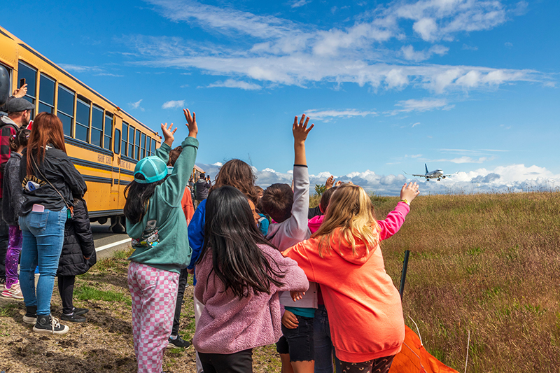 Children standing near a school bus raise their arms as a plane approaches to land over an open grassy field under a partly cloudy sky.