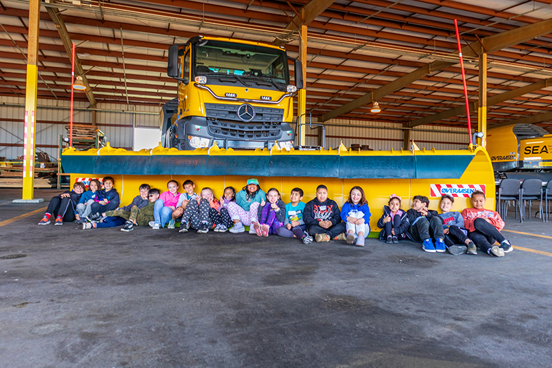 Group of children seated in a row on the floor of a maintenance hangar in front of a large yellow snowplow truck.