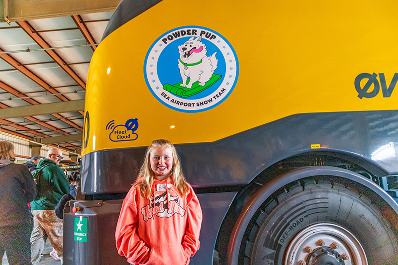 Child standing in front of a large yellow snow‑removal vehicle inside a hangar, with the vehicle’s ‘Powder Pup’ SEA Snow Team logo visible.