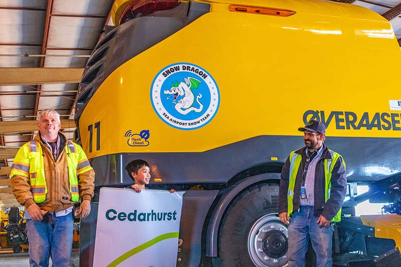 Two airport staff members and a child stand beside a large yellow snow‑removal vehicle inside a hangar, with the vehicle’s ‘Snow Dragon’ SEA Snow Team logo visible.