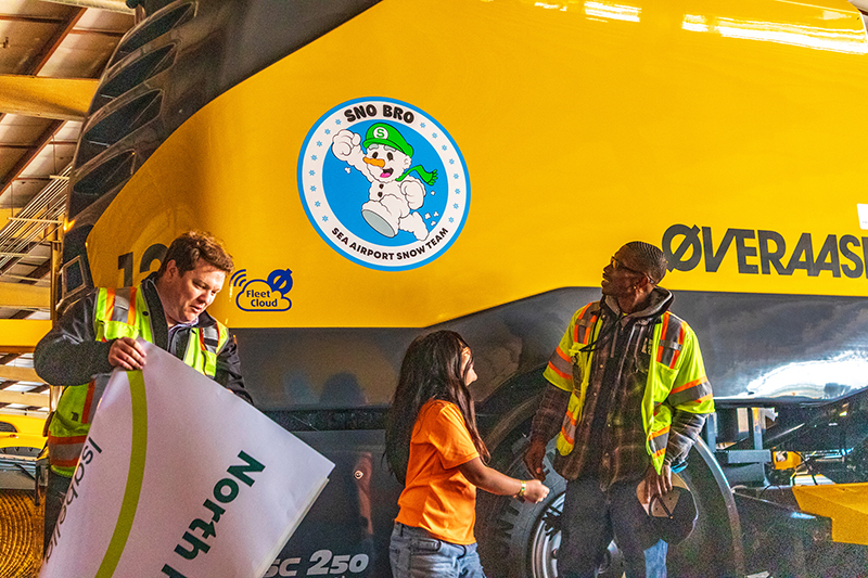 Two airport staff members and a child stand beside a large yellow snow‑removal vehicle inside a hangar, with the vehicle’s ‘Sno Bro’ SEA Snow Team logo visible.