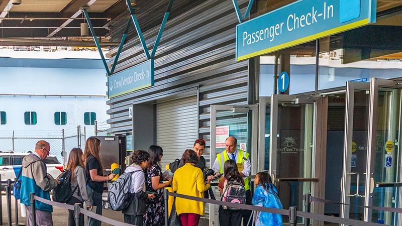 People line up at a passenger check‑in area where staff assist travelers near a terminal entrance.