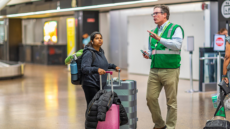An airport customer service worker in a green vest speaks with a traveler who is standing with rolling luggage.