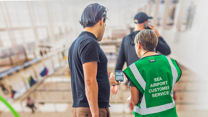 An airport customer service worker in a green vest assists a traveler while standing on an upper level overlooking the terminal.