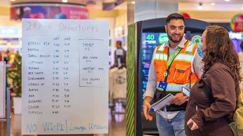 Two people talk near a handwritten departures board inside an airport, with one person wearing a safety vest.