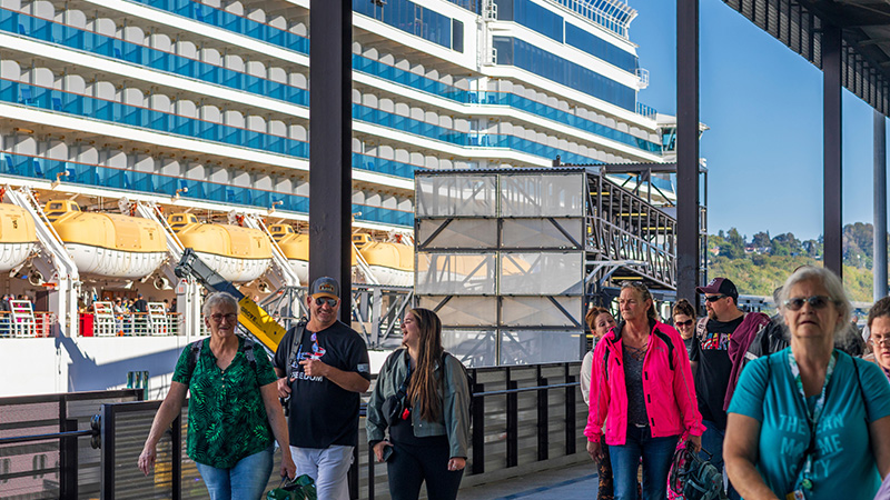 People walk along a covered terminal walkway beside a docked cruise ship with lifeboats visible.