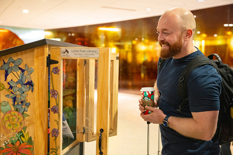 A person standing in an airport area looks at a Little Free Library decorated with colorful floral artwork while holding a drink and a piece of luggage.