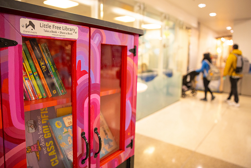 A brightly painted Little Free Library filled with children’s books sits in a hallway, with people walking in the background.