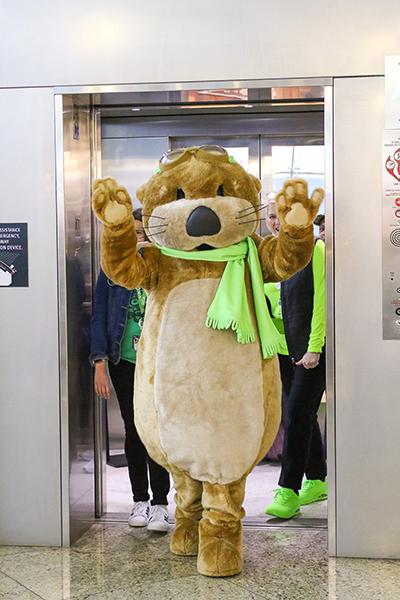 Jett, an otter-like mascot wearing goggles and a green scarf, steps out of an elevator with raised arms while several individuals stand behind.