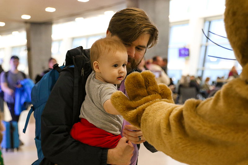 An adult holds a small child who reaches out to touch the raised paw of Jett, an otter-like mascot, in a busy indoor setting.