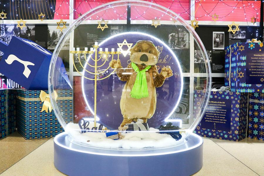 JETT the SEA Airport mascot stands inside a large snow‑globe display decorated with Hanukkah-themed elements, including a menorah, dreidels, and star designs.