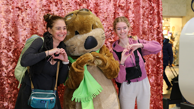 Two individuals pose with Jett, an otter-like mascot wearing goggles and a green scarf, all making heart shapes with their hands in front of a pink backdrop.