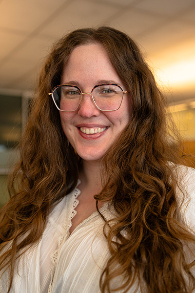 Trudy wearing glasses and a light-colored top is standing in an office environment, looking toward the camera.
