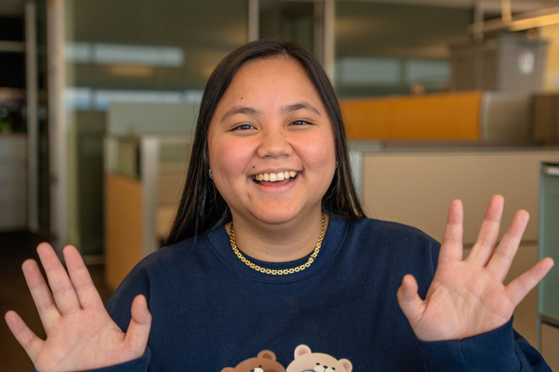 Dianne wearing a dark sweatshirt is in an office environment with both hands raised toward the camera.