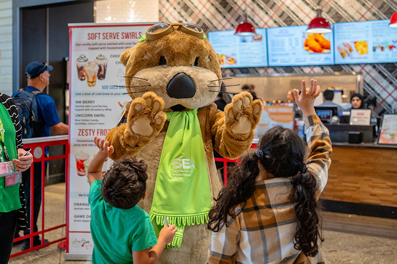 Two children greet JETT, a large otter mascot wearing a green SEA Airport scarf, inside a food court area.
