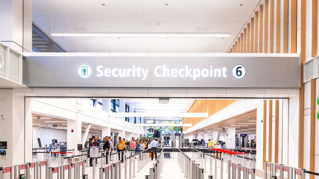 The entrance to Security Checkpoint 6 at SEA Airport is shown with a large overhead sign and queuing lanes leading to the screening area where travelers and staff are visible.