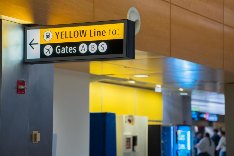 Overhead airport sign with a yellow header reading ‘YELLOW Line to:’ followed by directional symbols and text pointing left toward Gates A, B, and S. The sign hangs from a ceiling within a brightly lit terminal area that includes yellow wall panels and various fixtures in the background.