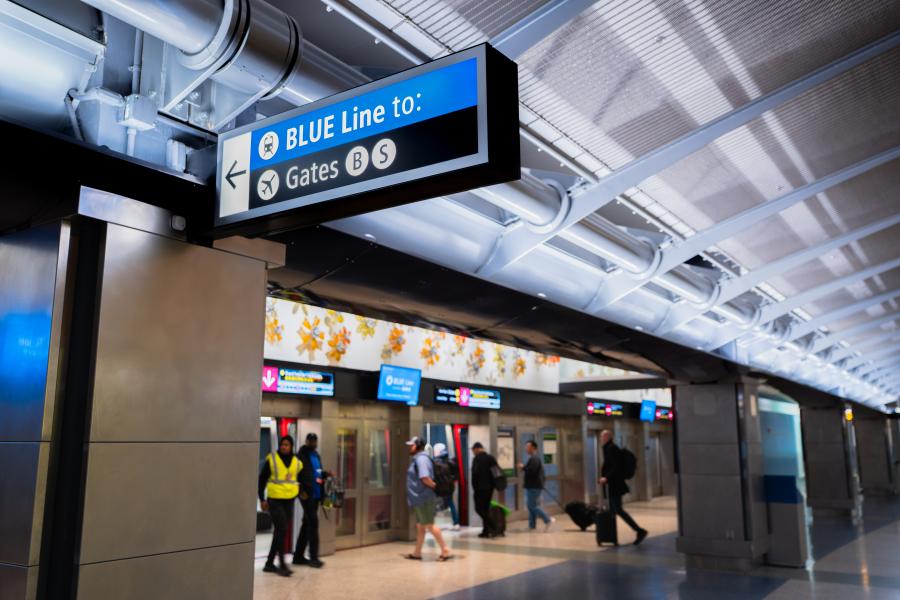 Overhead airport sign with a blue header reading ‘BLUE Line to:’ and directional symbols pointing left toward Gates B and S. The sign hangs beneath a ceiling with exposed structural beams and lighting. Below the sign, travelers move through a corridor lined with shuttle‑train doors, monitors, and wall decorations.