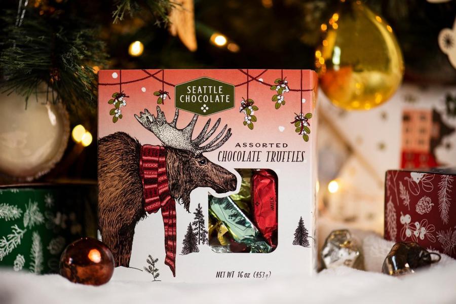 Box of Seattle Chocolate assorted truffles with an illustrated moose wearing a scarf, displayed under a Christmas tree with ornaments and lights.