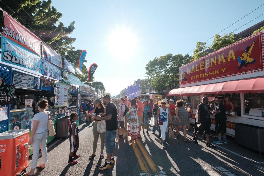Street festival scene with food booths on both sides of a sunlit road, including stands displaying signs for salmon dishes and piroshky. A crowd of people walks between the booths, browsing menus and ordering food, with trees and bright sunlight in the background.