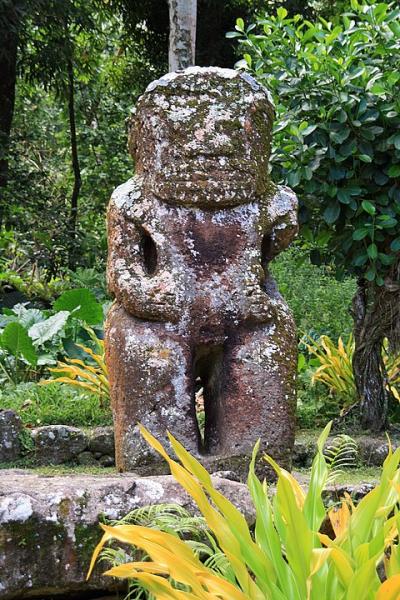 A weathered stone statue of a standing humanlike figure in an outdoor garden surrounded by greenery.