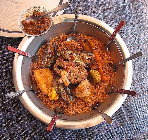 Large shared platter of Thieboujen, with seasoned rice topped with fish and assorted vegetables, surrounded by several spoons.