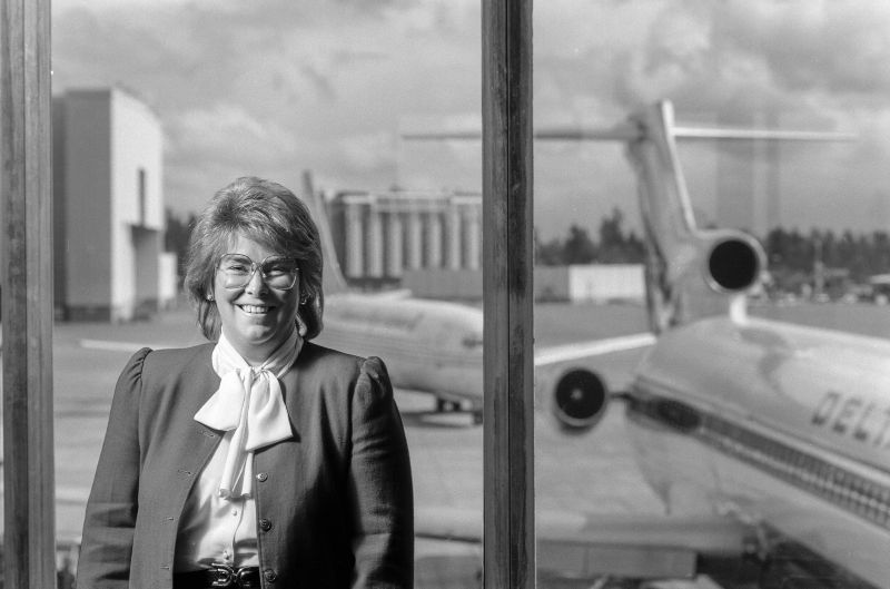 Andrea Riniker smiling near an airport window, with a Delta jet visible on the tarmac behind her. 