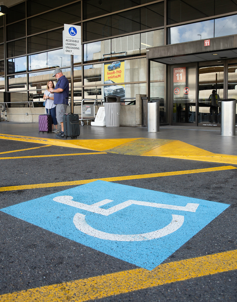 Accessible loading zone at an airport with a large blue accessibility symbol painted on the pavement and an entrance marked 'Door 11.