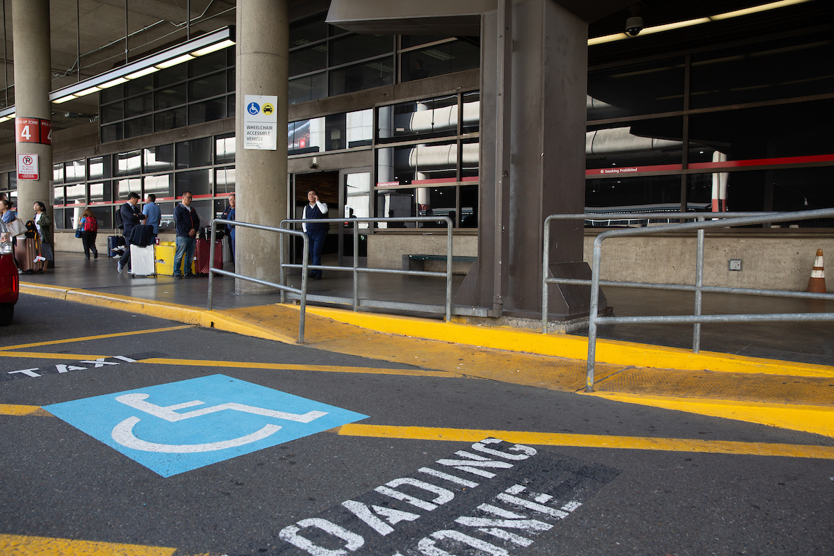 Accessible loading zone at an airport with a blue accessibility symbol on the pavement and a ramp with railings leading to the terminal.