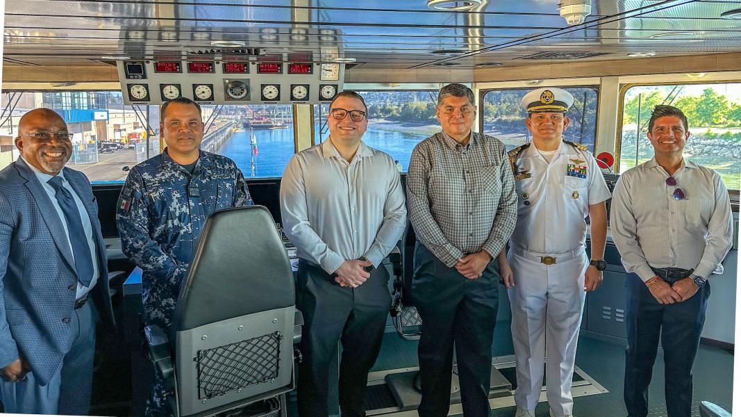 A group of people standing inside a ship’s bridge with control panels and windows showing the waterfront behind them.
