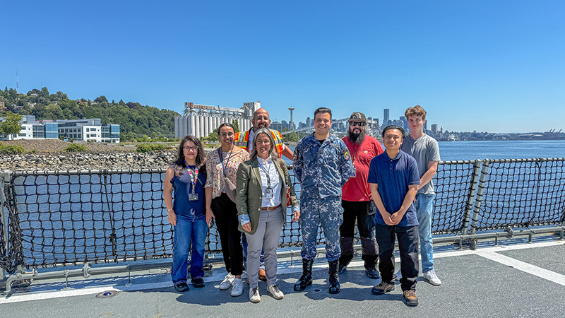 A group of people standing on a ship’s deck with a waterfront city skyline in the background.